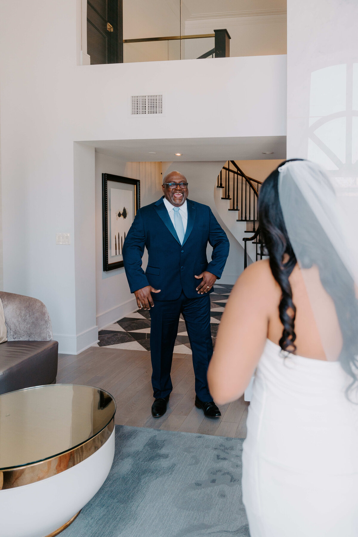 The image shows a man in a blue suit smiling warmly, standing in a modern living room. A woman in a wedding dress faces him, suggesting a joyful moment.
