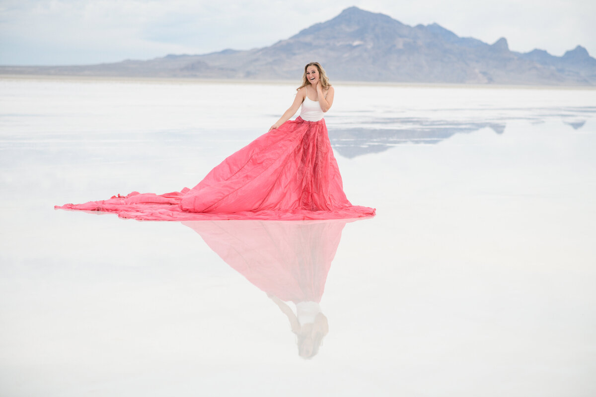 senior photos dresses with young woman in a large pink gown walking on salt flats with the mountains in the distance as her reflection is below her taken by denver senior photographer
