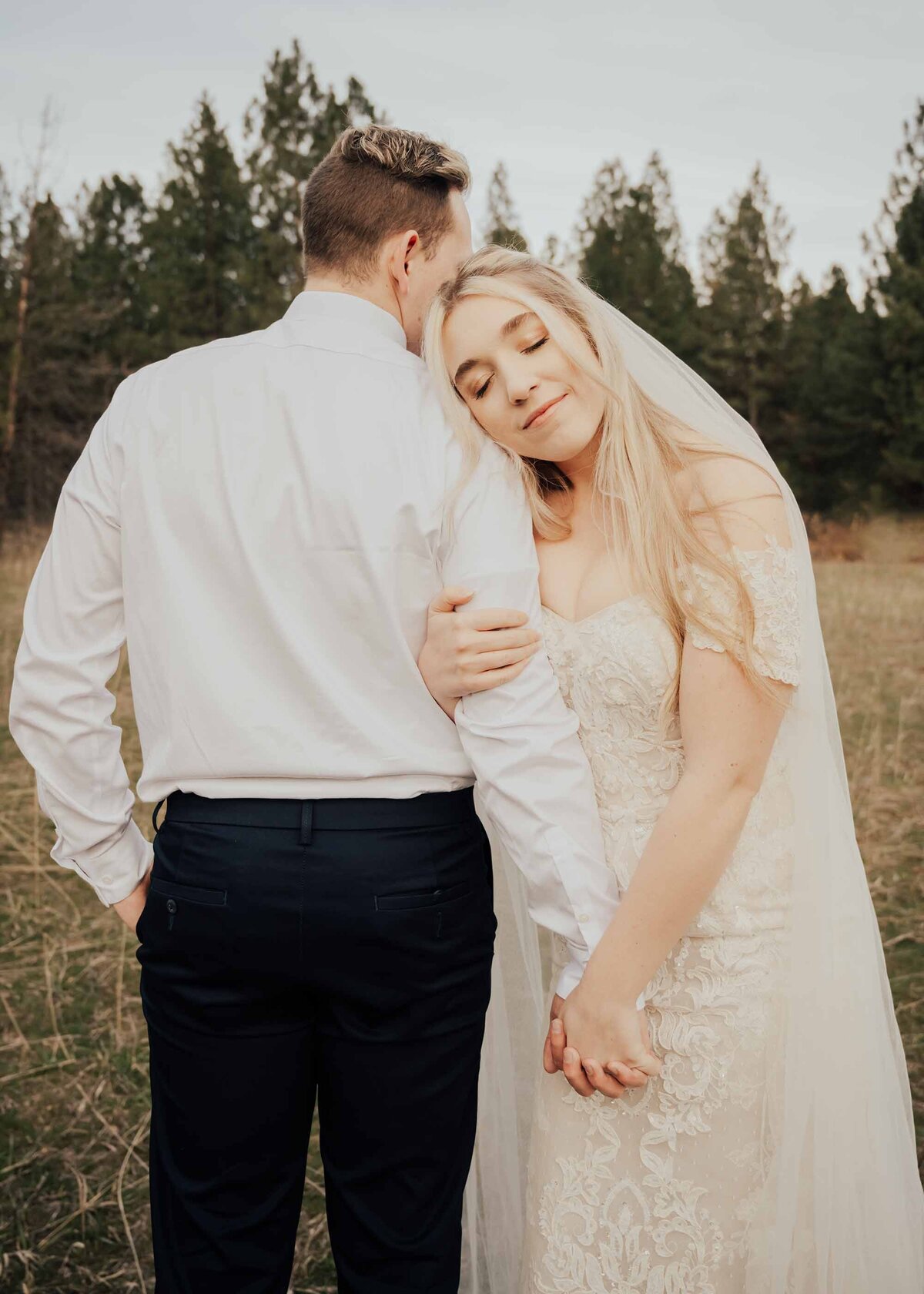 Maddie Rae Photography bride and groom standing side by side holding hands. he is facing away and she is facing the camera resting her head on his shoulder. he is resting his head on her head