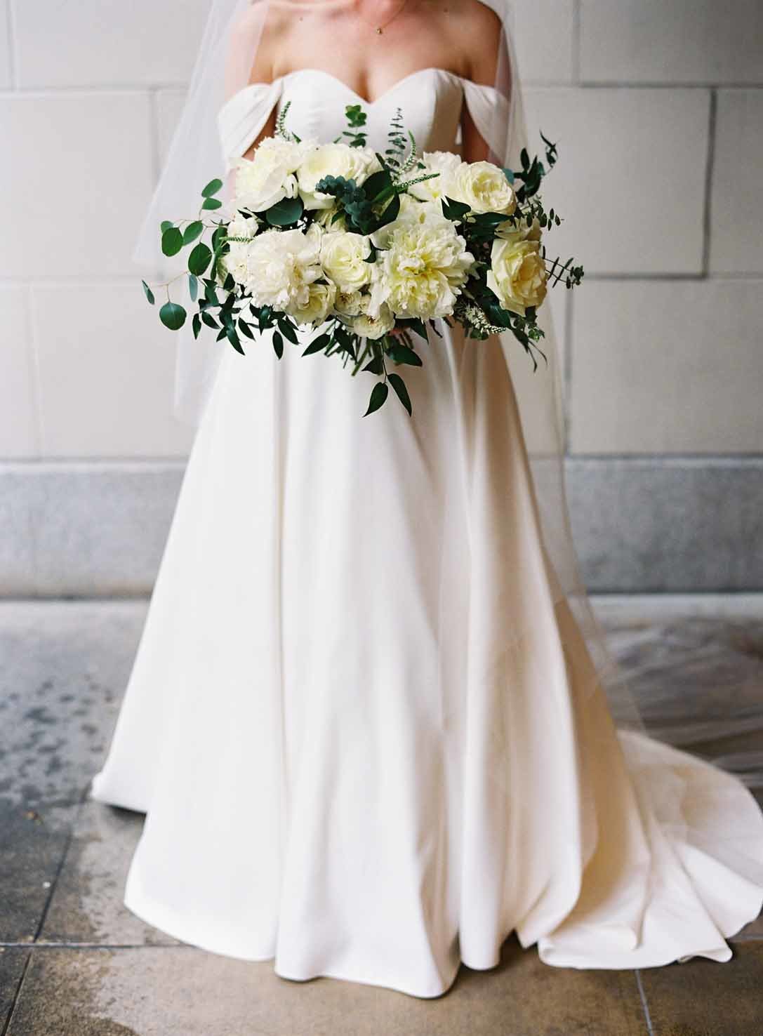 bride in long white dress holding an oversized bouquet of white roses and greenery