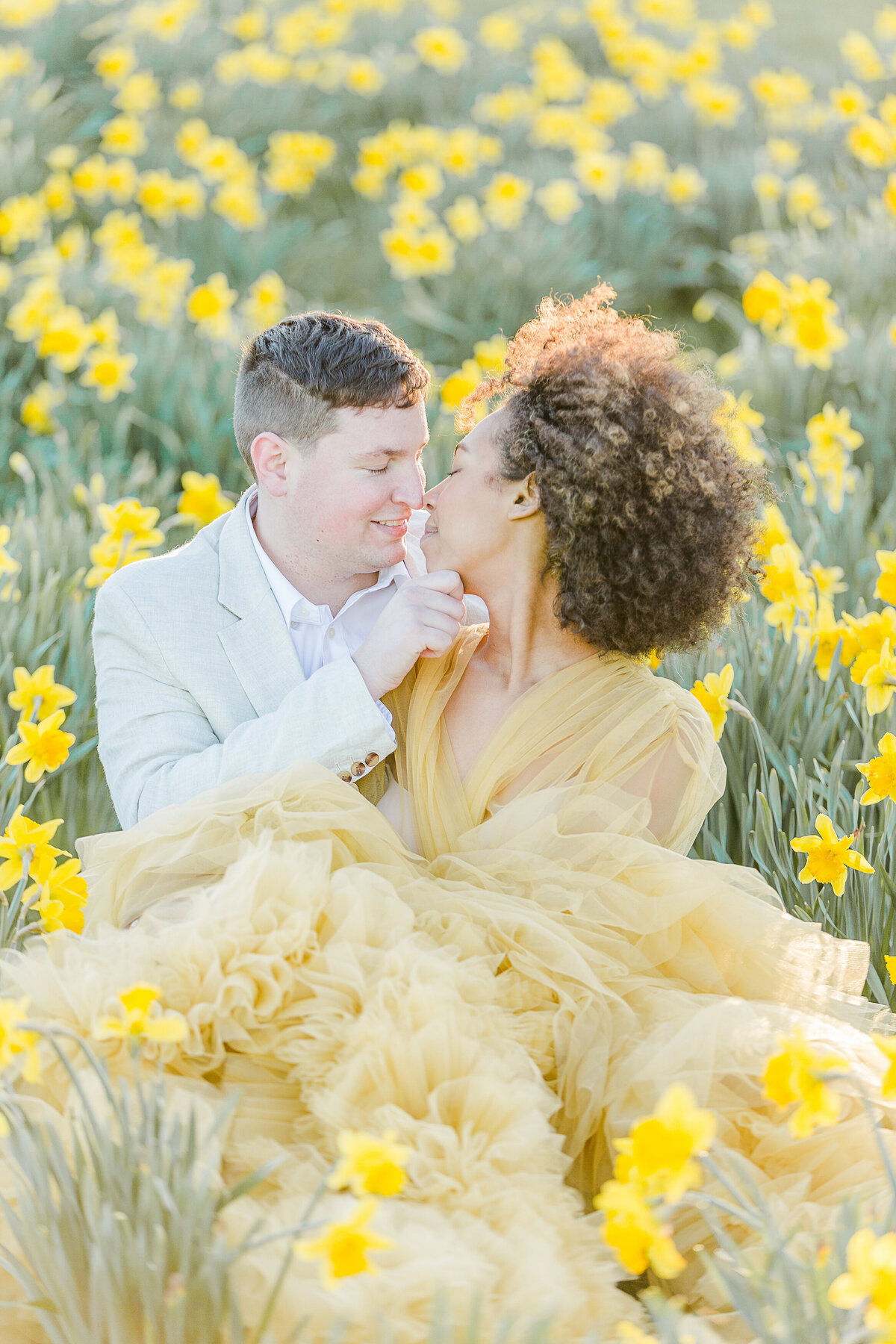 Man and woman are sitting amongst the daffodils at Salve Regina's Cliff Walk. The woman is sitting slightly in front of the man and is turning around to share a kiss. Captured by Rhode Island Wedding and Engagement Photographer Lia Rose Weddings