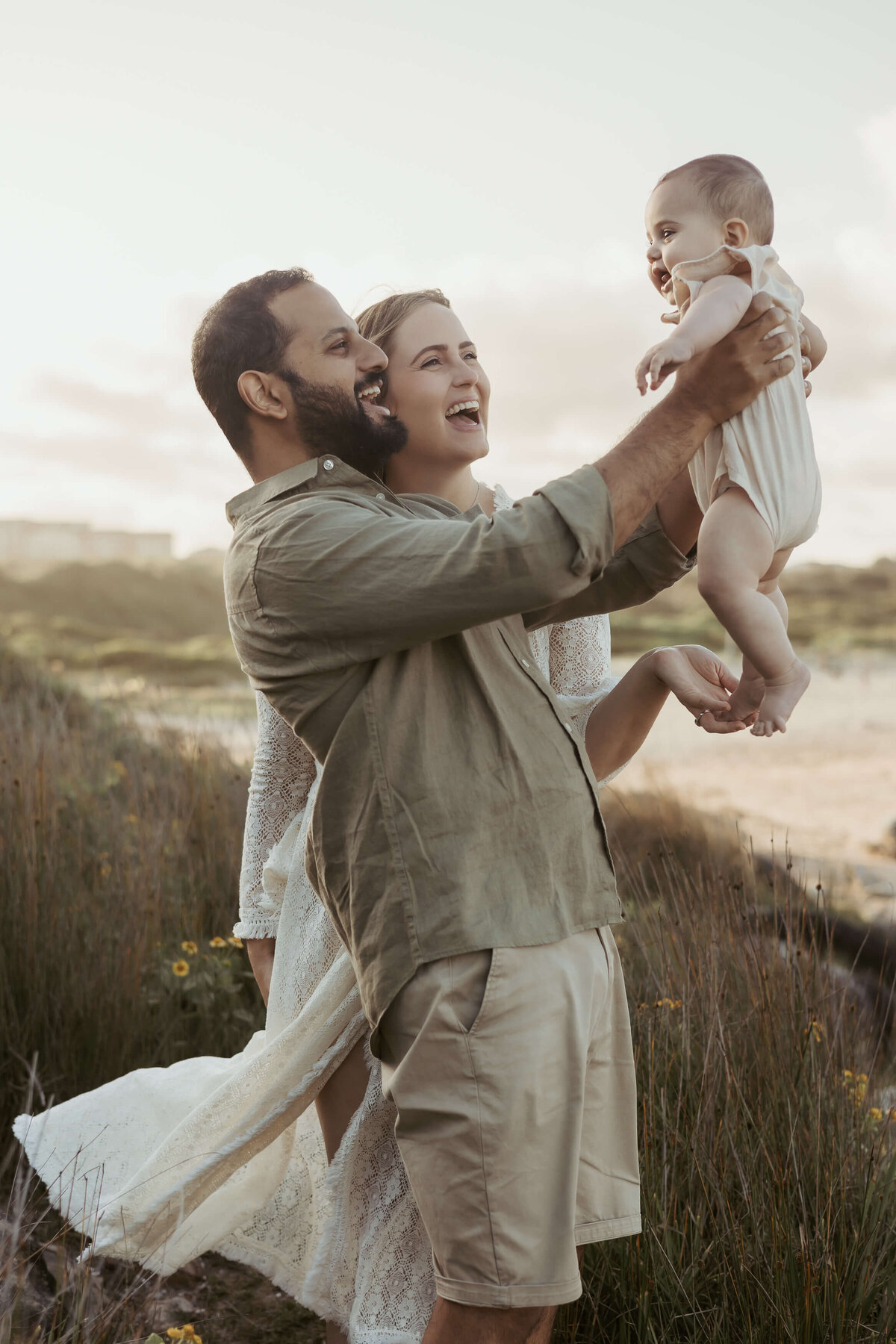 Dad in a green linen shirt and tan chino shorts is next to his wife are smiling widely while he holds up their giggling baby.