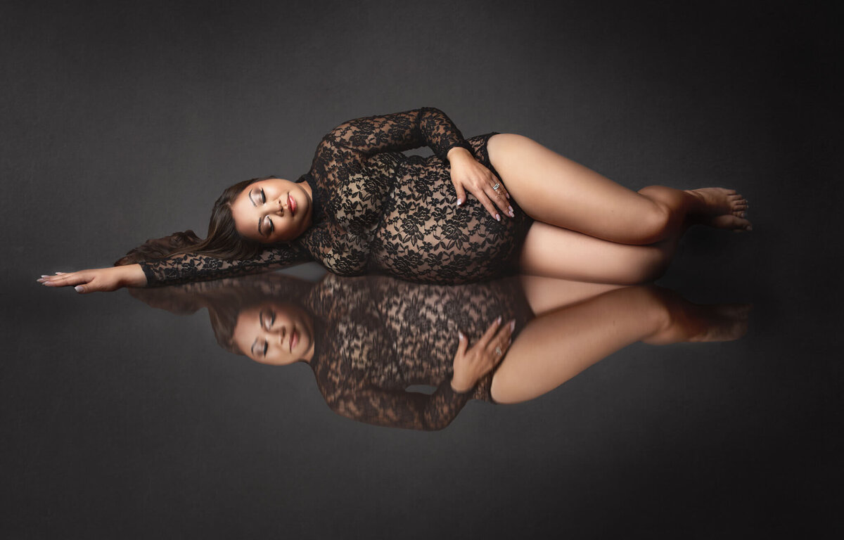 expecting mother laying on her side in black lace body suit with reflection below at hamilton, ON photography studio