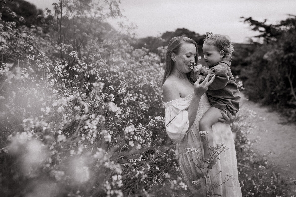 Pregnant mom holds toddler daughter on belly while smelling wildflowers