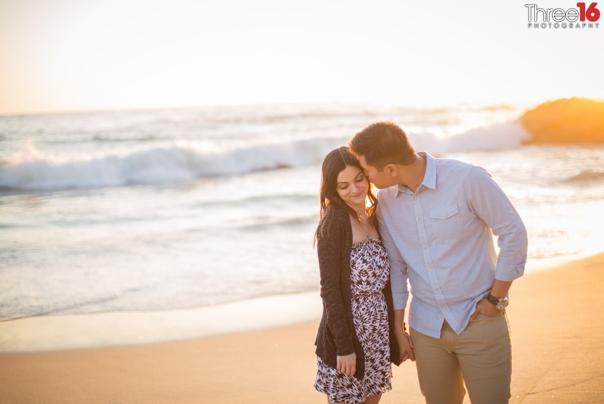 Groom to be leans over and kisses his fiance on the cheek with the ocean behind them