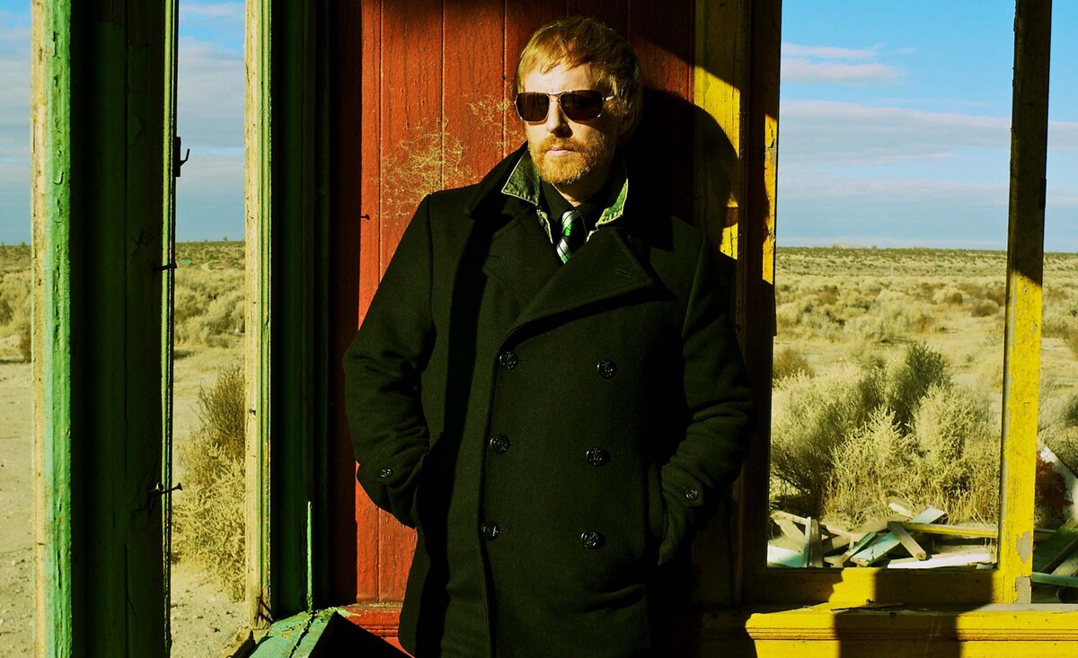 Male musician portrait Bob Kemmis wearing black overcoat with sunglasses leaning against open windows with desert sky background