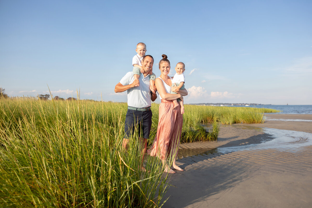 A modern, young family of four stands in tall green sea grasses along the Connecticut coastline during Golden Hour. Large homes and coming clouds dot the background.