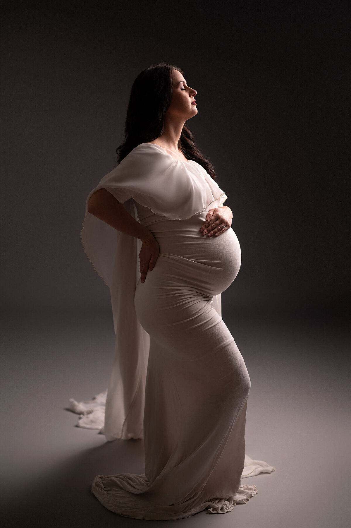 Full body shot of pregnant woman facing studio lighting in a floor-length, white, maternity gown.