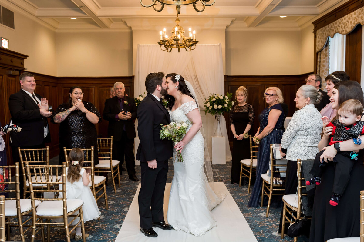 an Ottawa bride and groom kiss at the end of their Chateau Laurier wedding ceremony while family and guests clap and smile