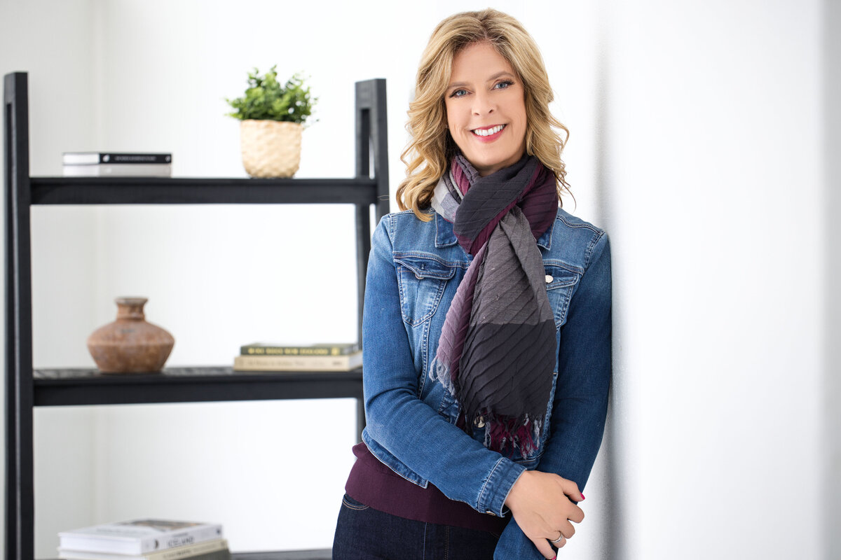 Professional headshot of a middle-aged Caucasian woman with blonde hair, captured in Cincinnati for personal branding. She is wearing a denim jacket and a stylish scarf, leaning casually against a white wall with a bookshelf in the background, adding a touch of personality and warmth. Perfect for updating LinkedIn profiles and personal branding materials. Optimize your Cincinnati headshots with expert photography services for a polished, professional image.