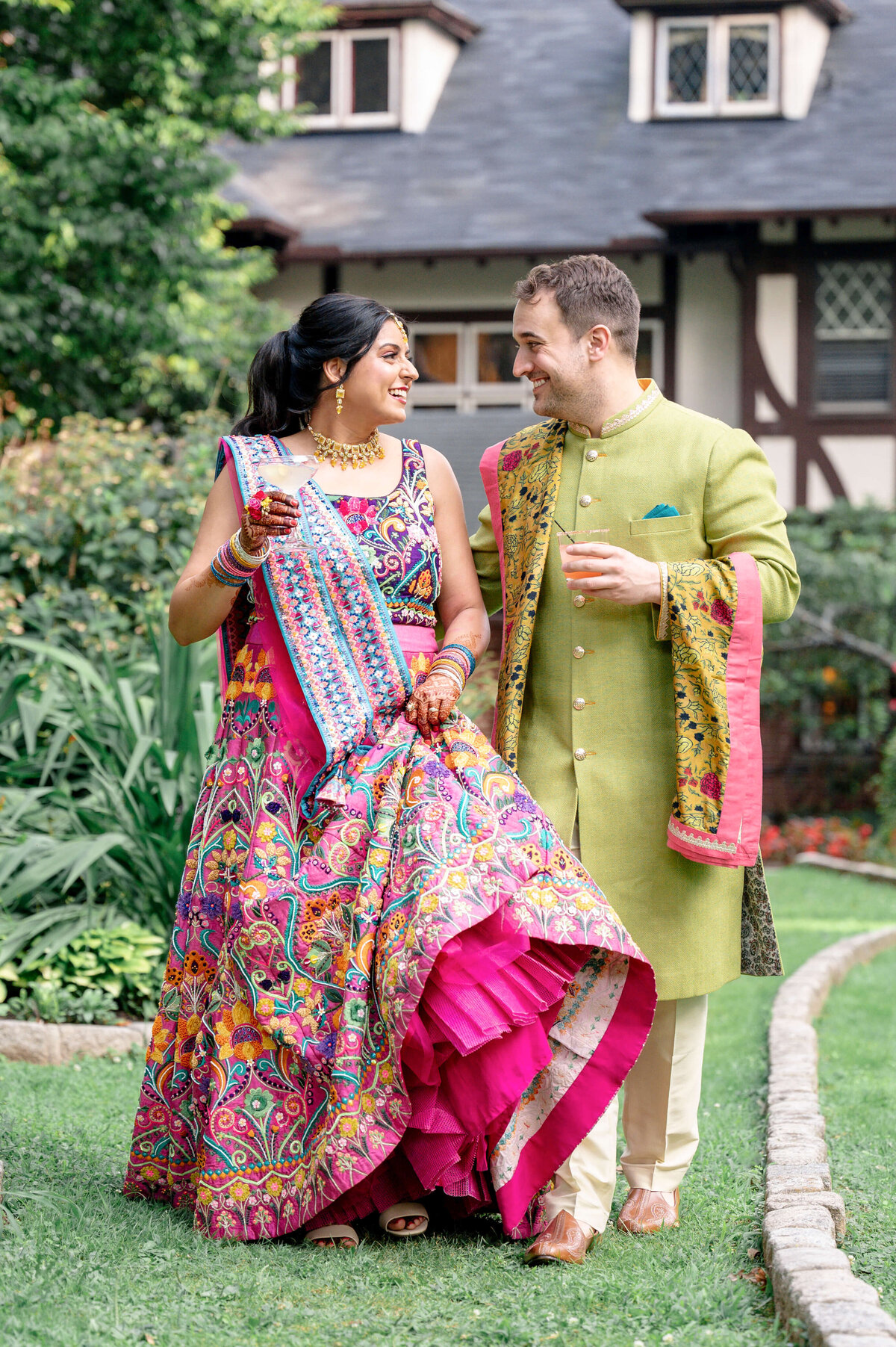 A couple is walking on grass in a garden. The woman wears a colorful, patterned lehenga, and the man is in a lime green sherwani. Both are smiling and looking at each other. There is greenery and a house in the background.