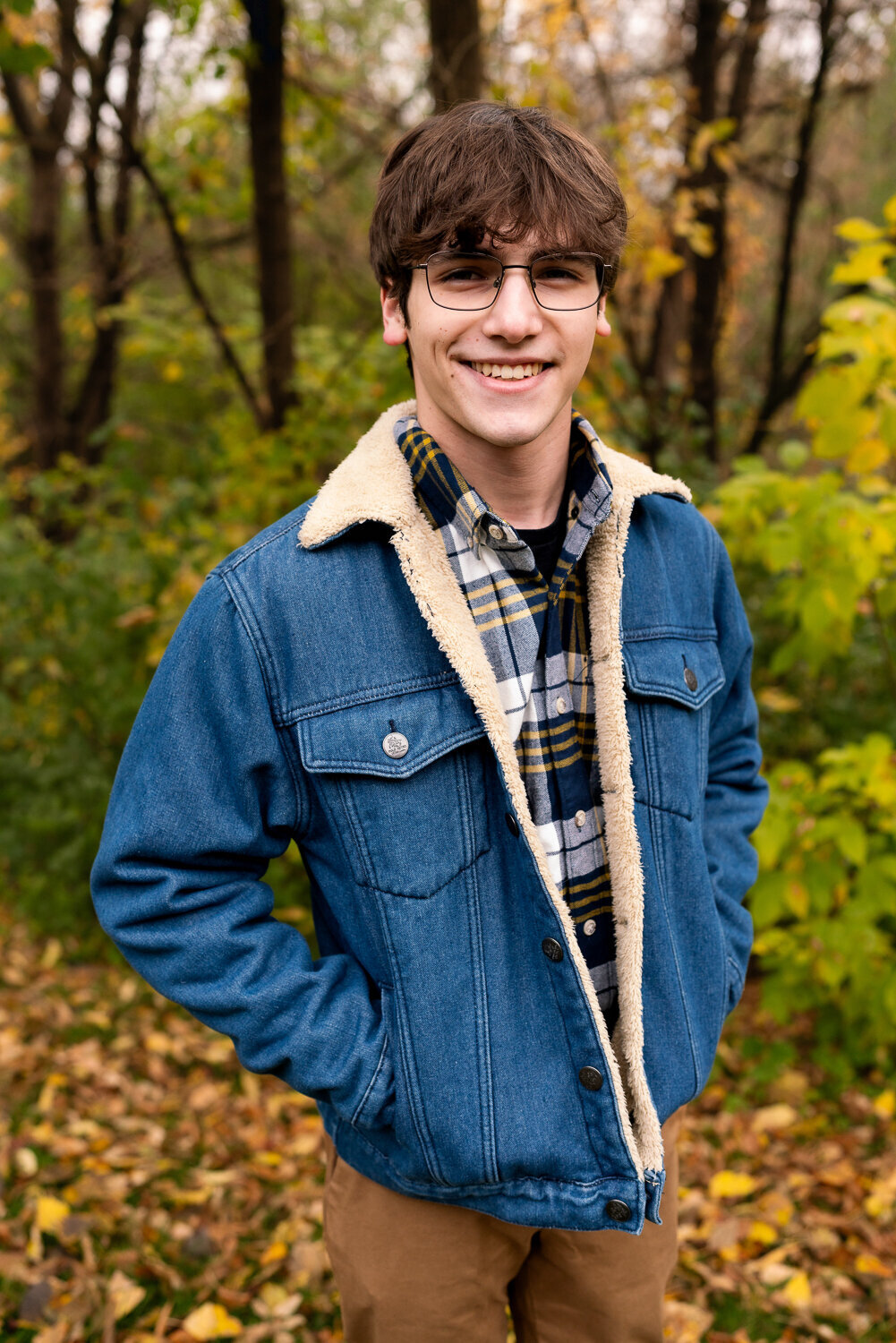 Senior boy with glasses dressed in a denim jacket smiles in front of the woods.