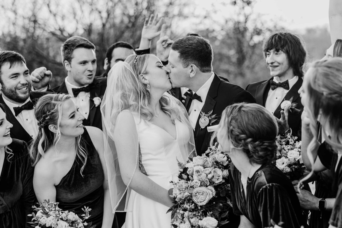 Bride and groom kissing at a park farm winery wedding surrounded by joyful guests in formal attire, captured in a black and white photograph.