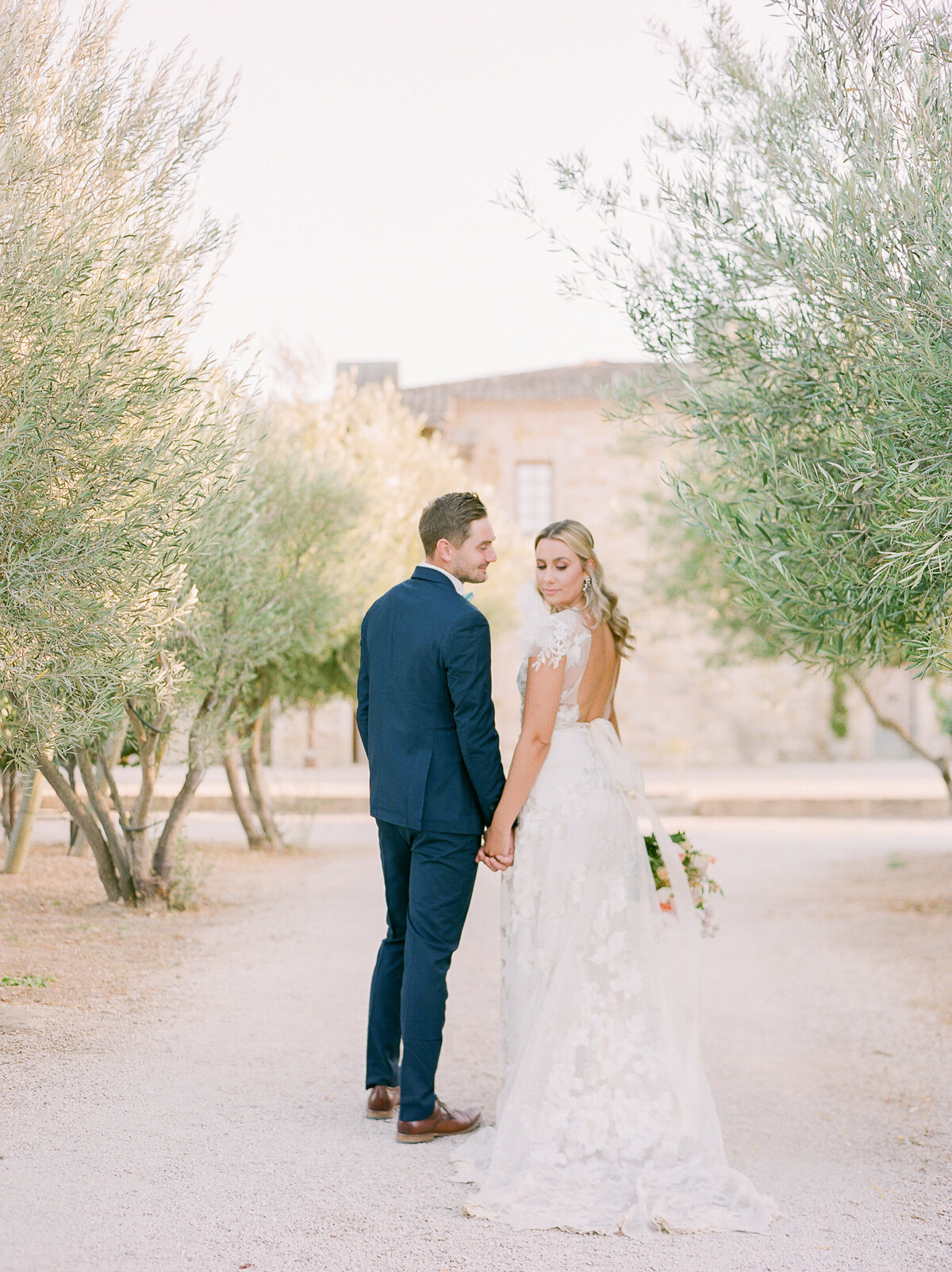 a bride and groom holding hands and walking away from the camera and looking at each other in a field of olive trees in  italy