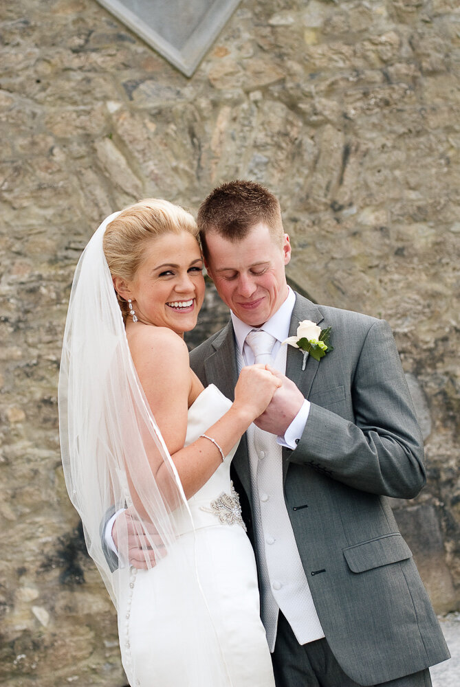 blonde bride wearing a mermaid dress and long veil embracing her husband who is wearing a light grey suit and white tie in front of Ross Castle in Killarney