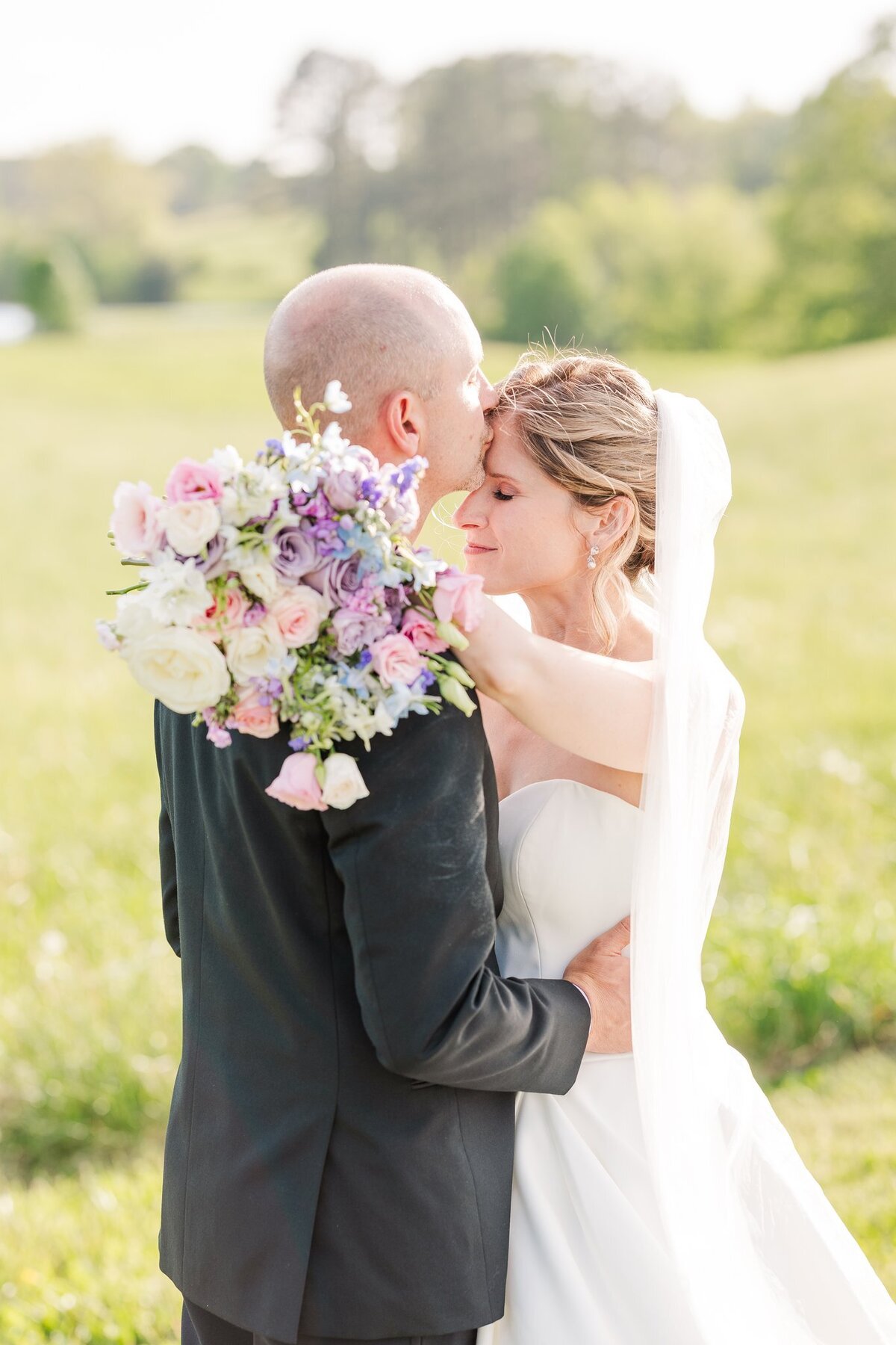 Groom kissing his wife on her forehead while she drapes her arms around him while holding her lavender and blush pink themed wedding bouquet