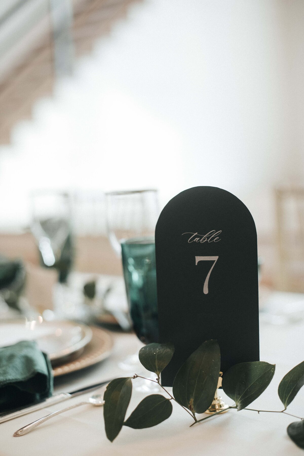 Rounded dark green place card with ivory cursive font on a gold place card holder’ set atop reception table.
