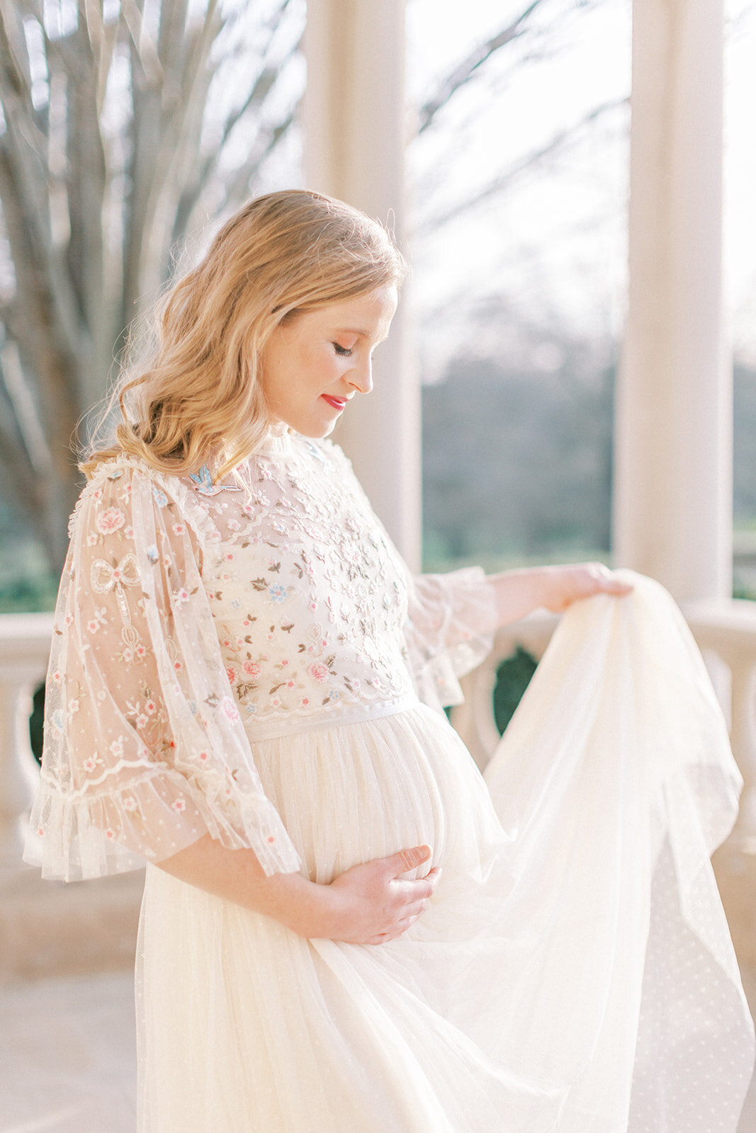 Pregnant blonde mother in Needle & Thread dress places on hand below her belly and the other hand holding up part of the dress.