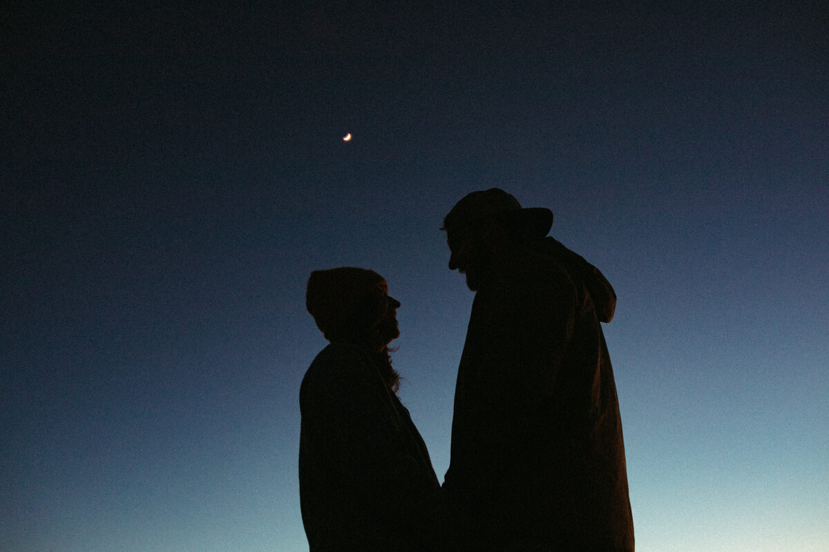 Silhouette of a couple standing facing each other with the night sky and the moon behind them at blue hour