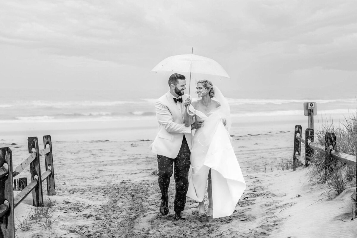 Bride and groom, walking off the beach, holding an umbrella during a rainstorm before going to their wedding reception at the Reeds in Stone Harbor