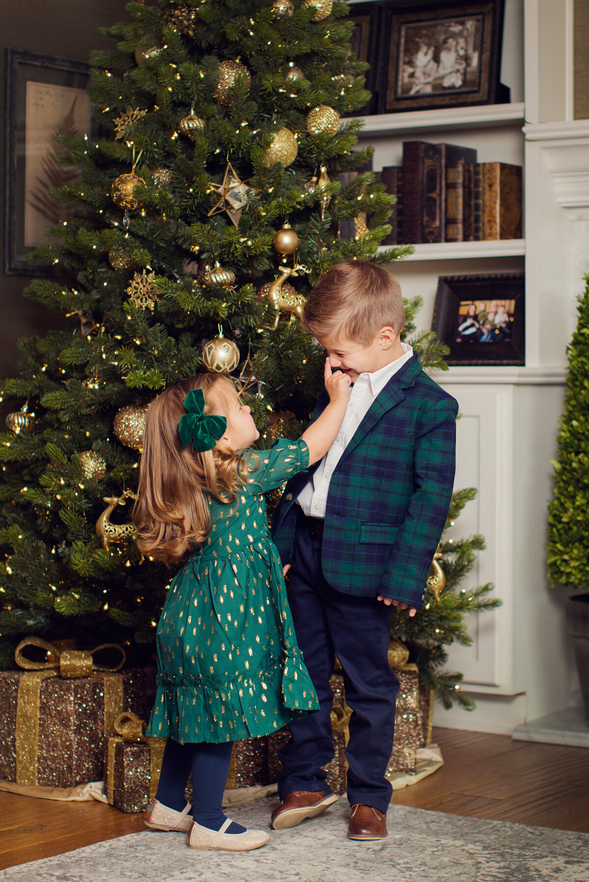It's Christmas time and the tree is up. Kids dressed up in blue and green, she is touching her brother's nose gently, he is laughing at her.