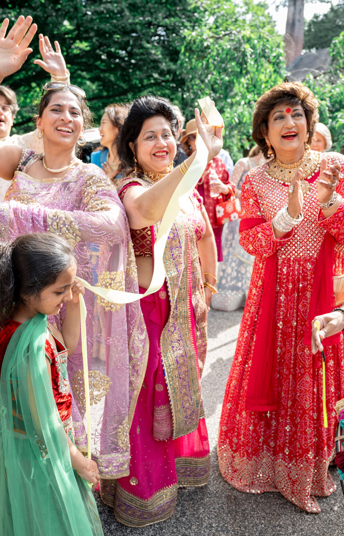 A joyful group of women, dressed in vibrant traditional attire, celebrate outdoors. They are smiling, clapping, and waving ribbons, with trees visible in the background, creating a lively and festive atmosphere.