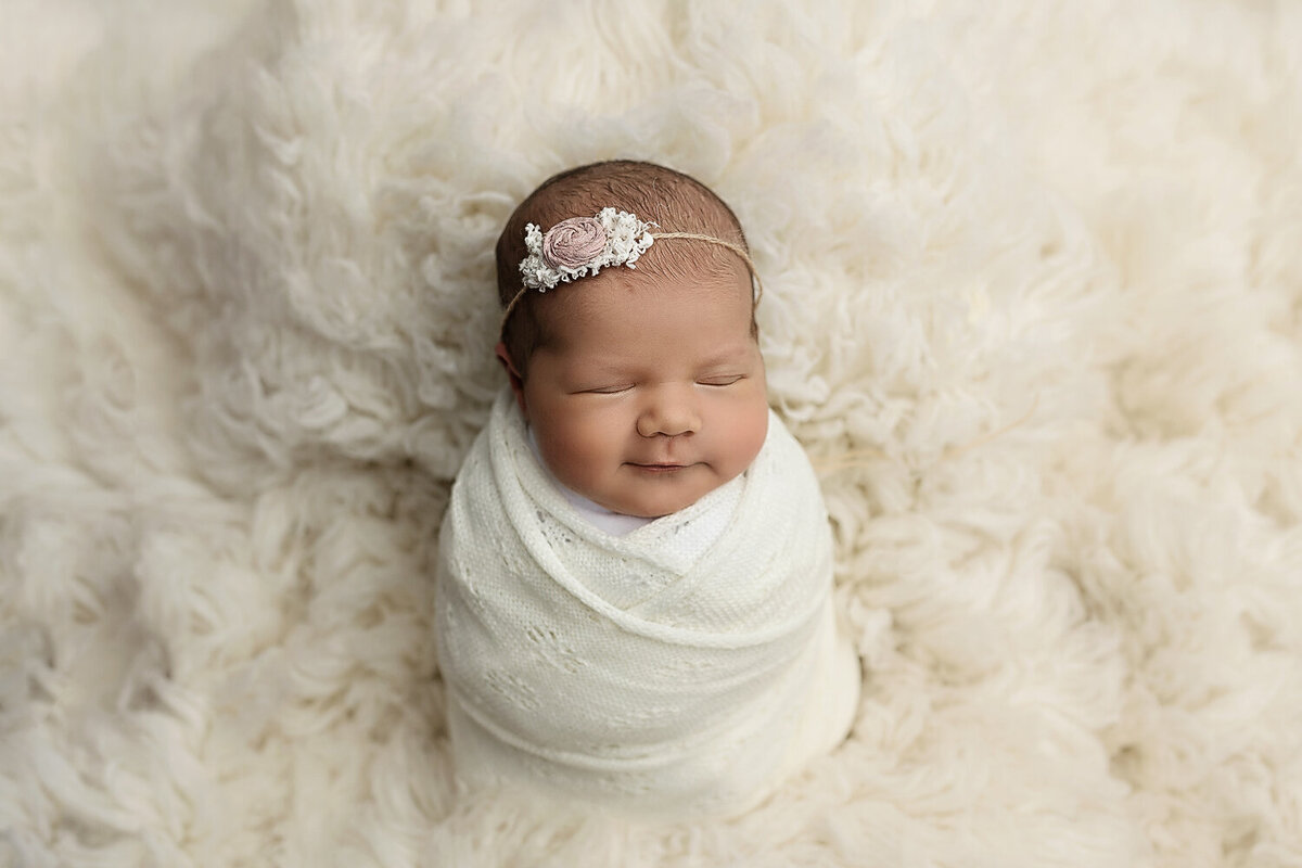A smiling newborn baby sleeps in a white swaddle in a studio
