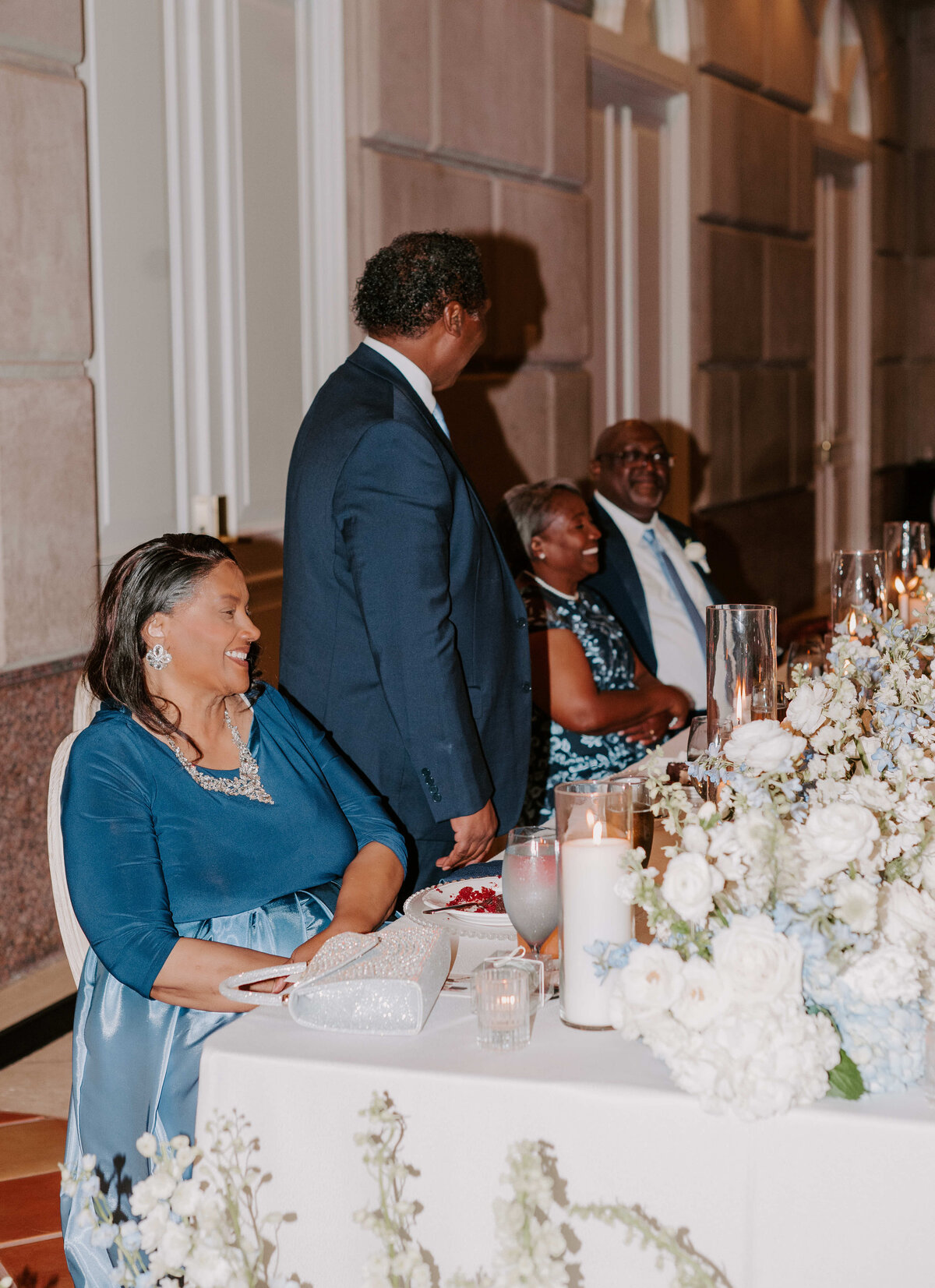 A joyful group at a wedding reception. The setting features elegant floral arrangements and candles on a table, with everyone dressed in formal attire.