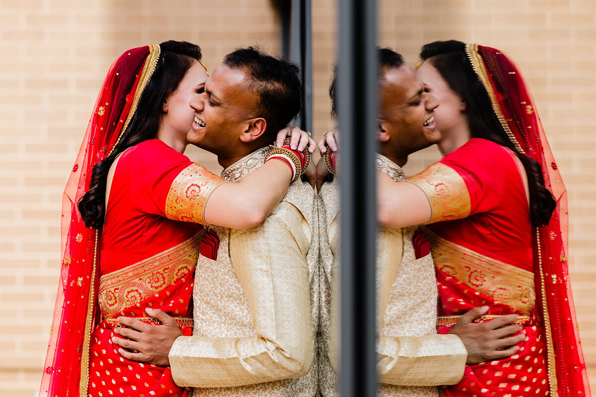 A couple embracing and laughing outside, with the bride in a red saree and the groom in a cream sherwani.