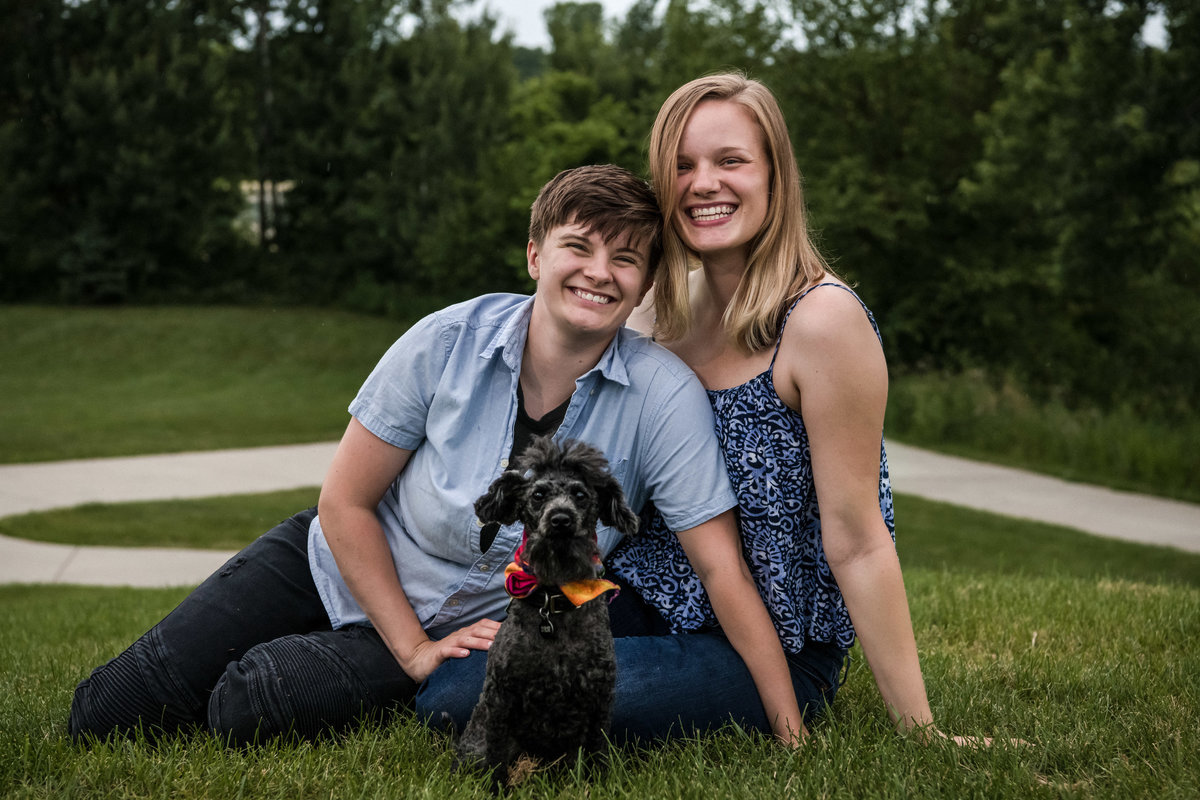 Lesbian couple smile while sitting in the grass with their Doodle puppy.