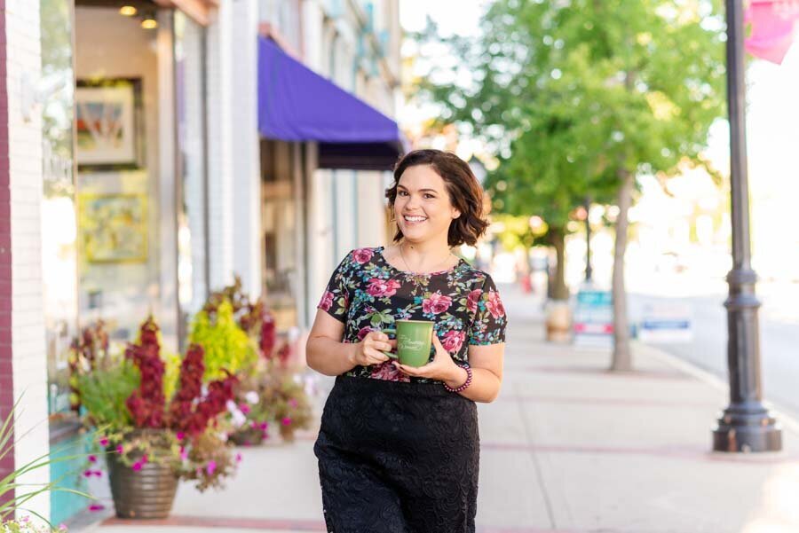 Woman smiling, holding a green mug, walking past storefronts on a sunny street lined with plants.