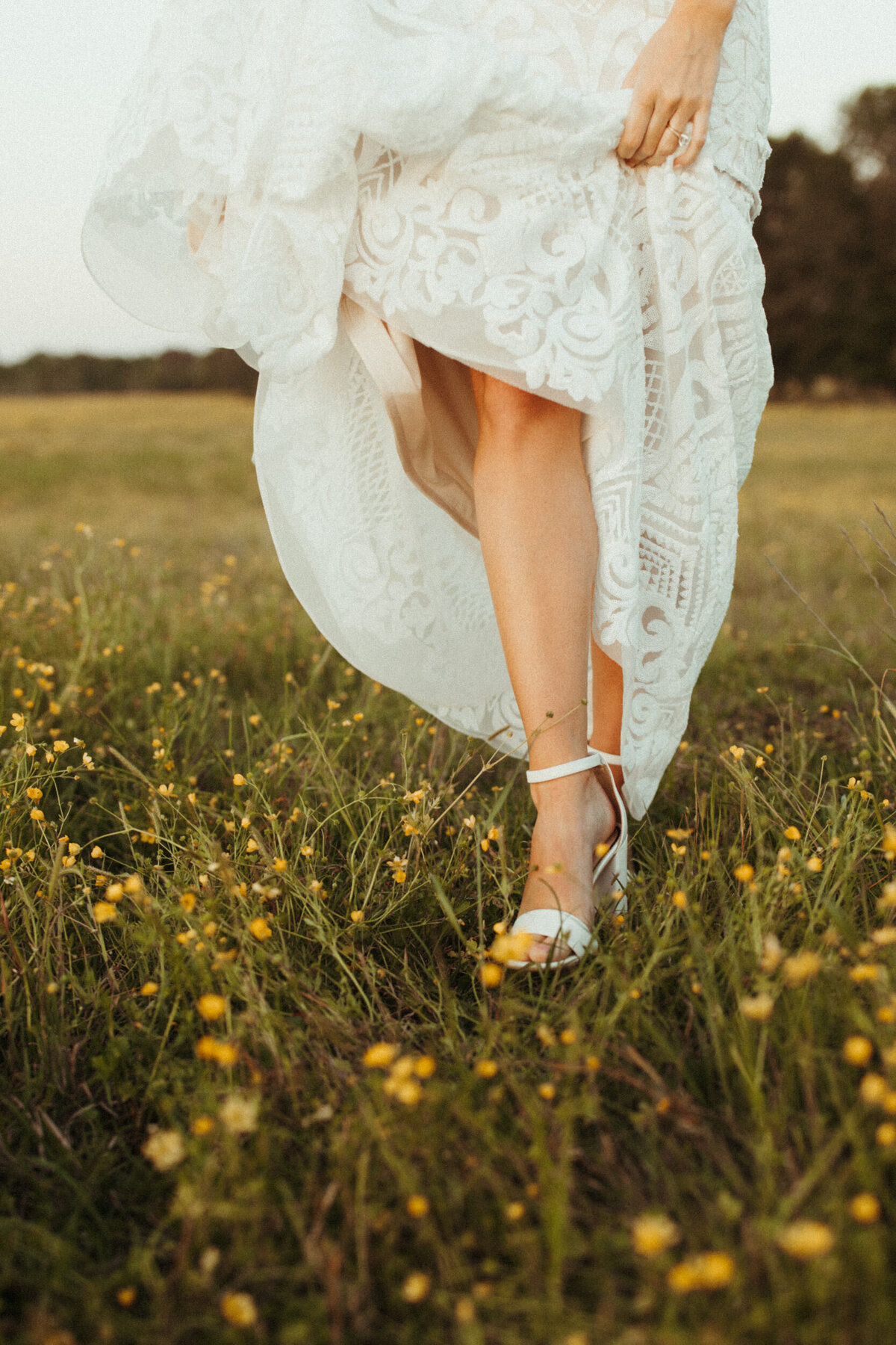 Bride holding up her wedding dress showing off her shoes as she walks through a field of wildflowers