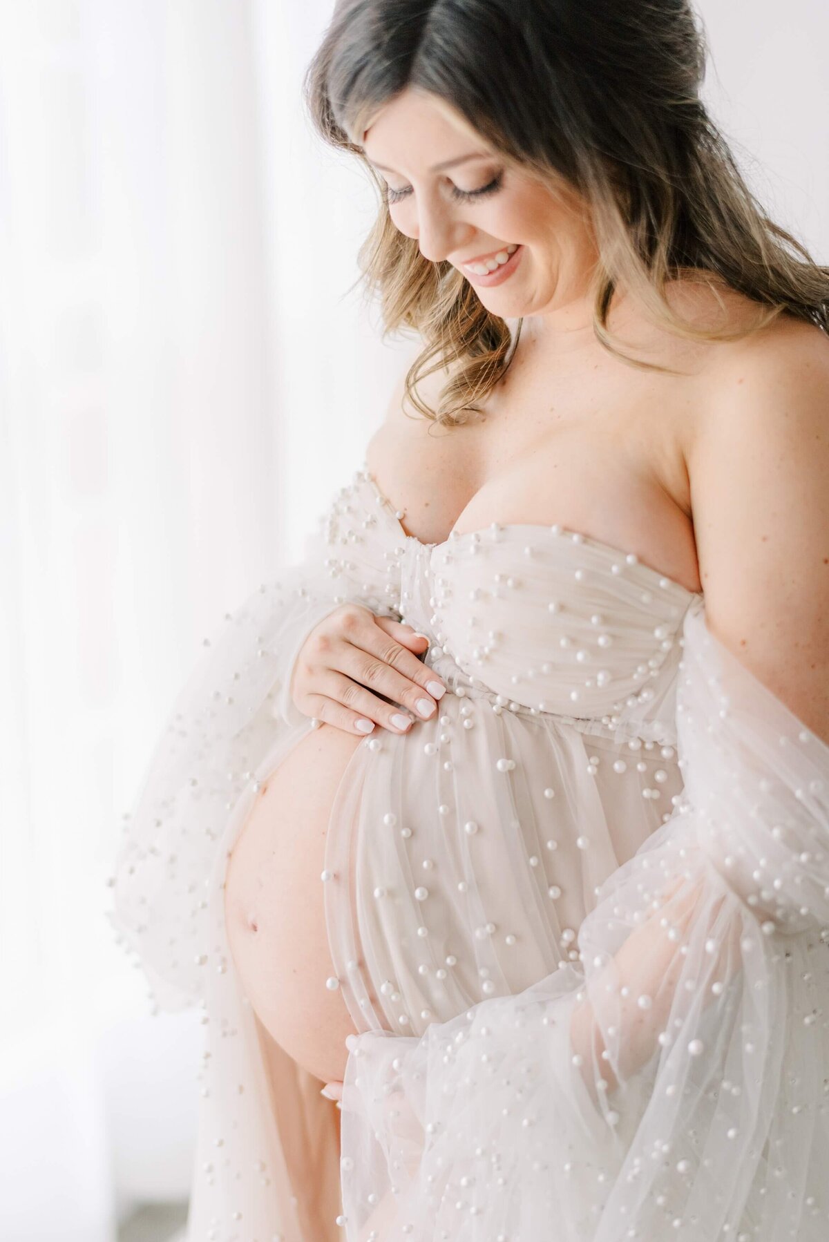 Pregnant woman looks down at her belly peeking out from a tulle gown with pearls