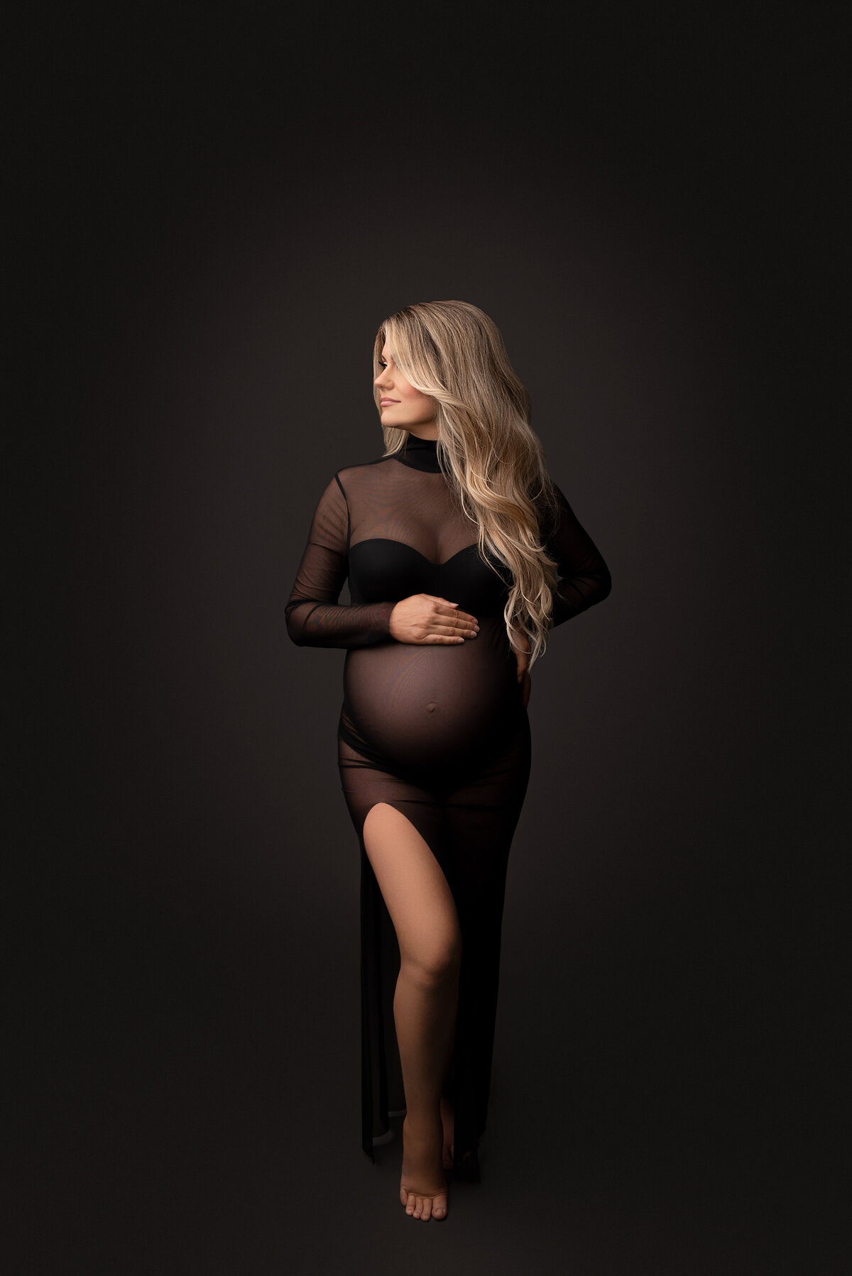 New Jersey's best maternity photographer Katie Marshall captures expectant mom for fine art maternity photos. Expectant mom in a sheer maternity photoshoot dress stands with her body facing the camera for a studio maternity portrait. Her knee is bent and peeks out from the slit in the dress. She is looking over her shoulder toward the light source.
