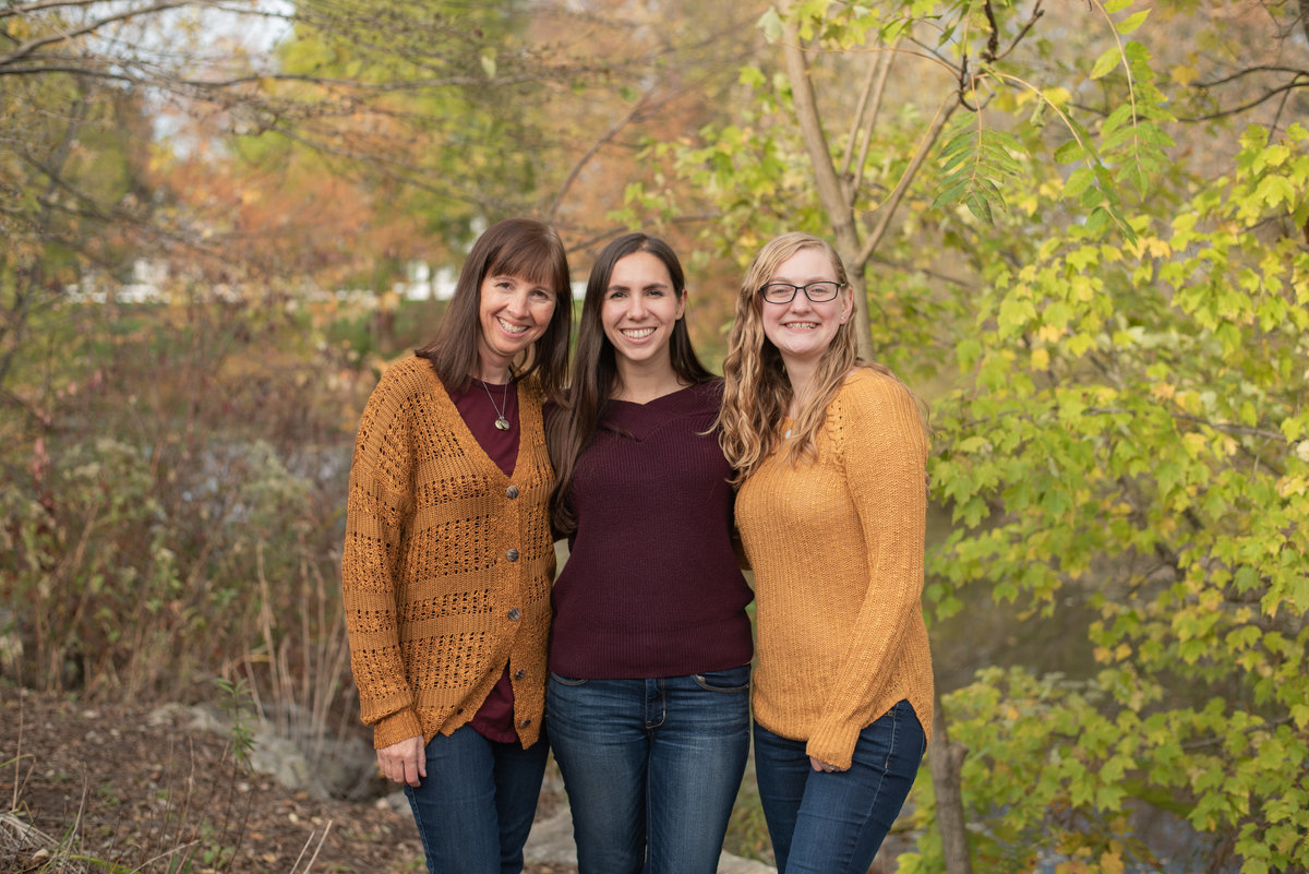 Mom and two Daughters in fall colors standing in front of tree-rimmed pond
