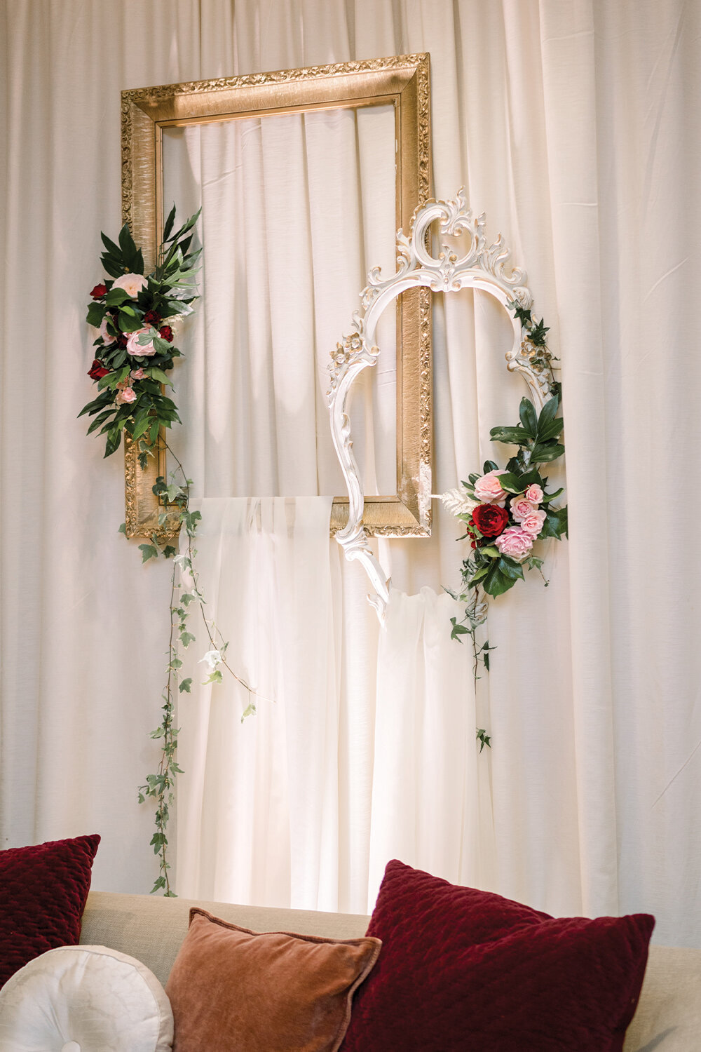 Ornate white and gold frames adorned with cascading burgundy and blush florals against sheer white drapes with uplighting behind the lounge area at a 50th wedding anniversary celebration.