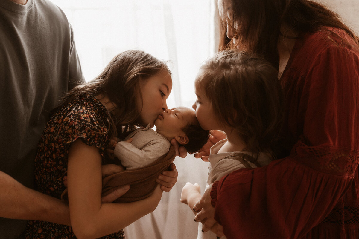 Two older sisters kissing on their newborn baby brother while their parents hold them.