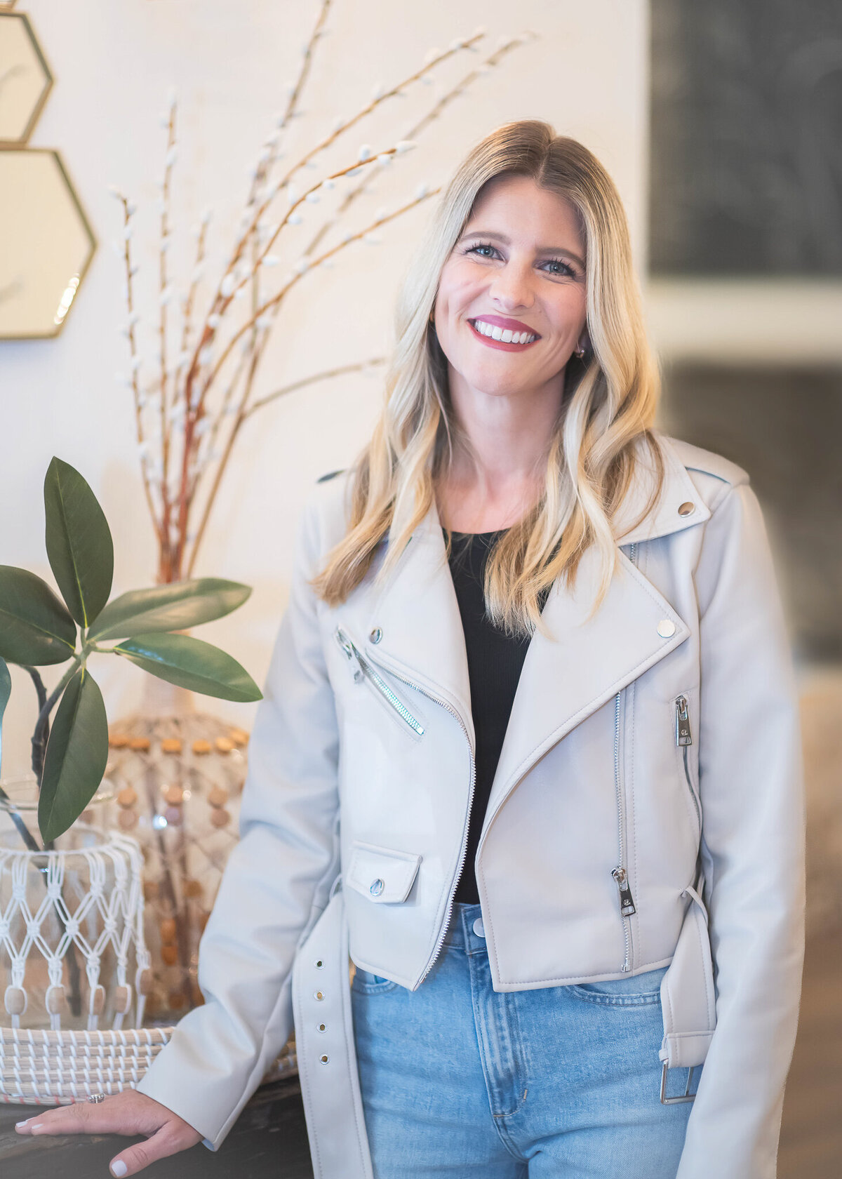 Lovely blonde woman wearing a light grey leathers jacket standing next to plants in vases on a table