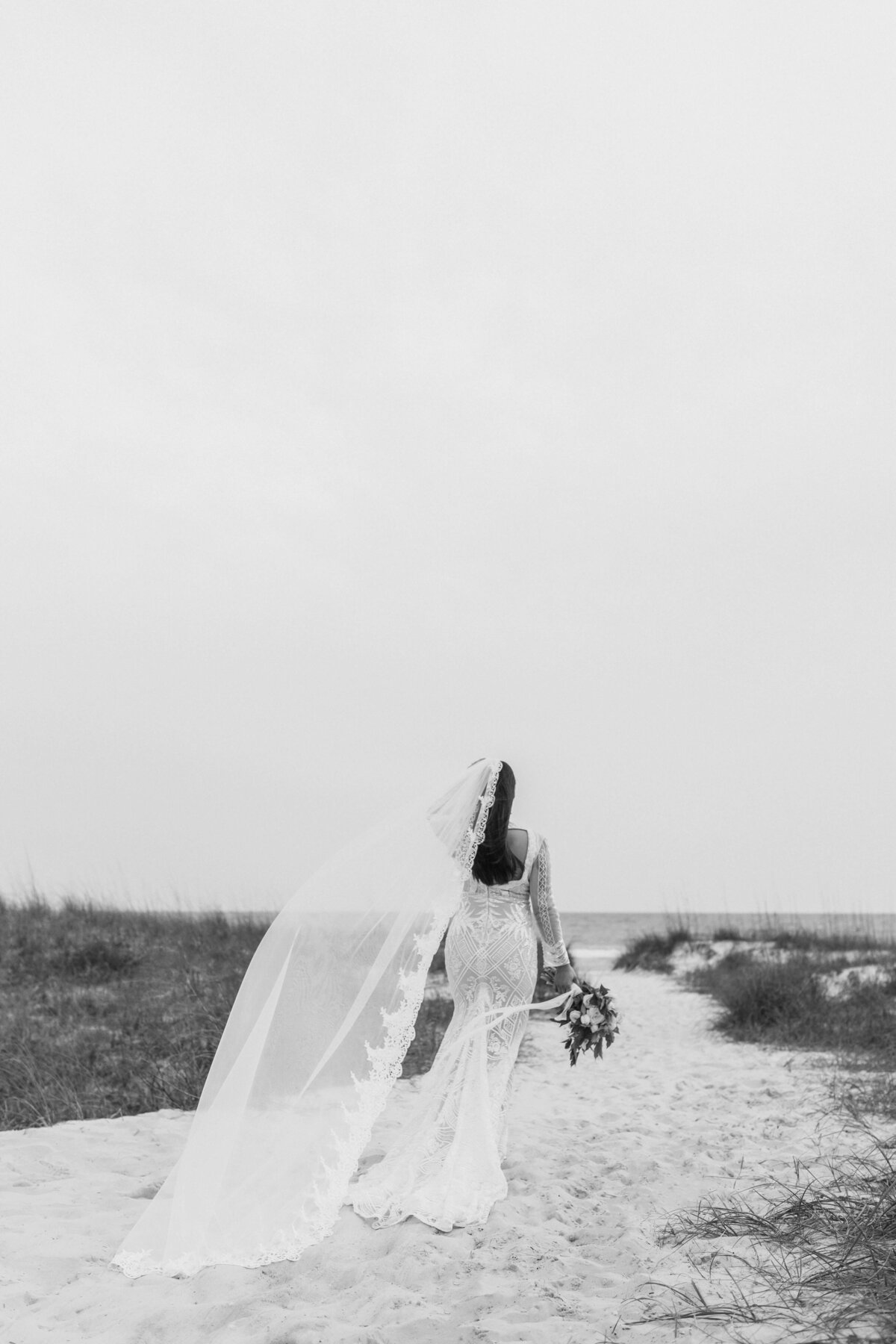 B&W shot of asian bride in bridal dress and veil walks on beach path - taken by panama city fl photographer Brittney Stanley