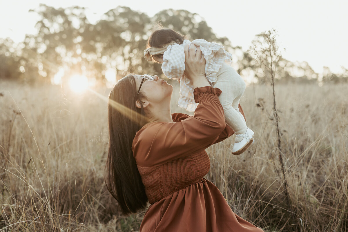 Mother in a grassy field is holding up her baby daughter to her face in the air while the sun glow behind them.
