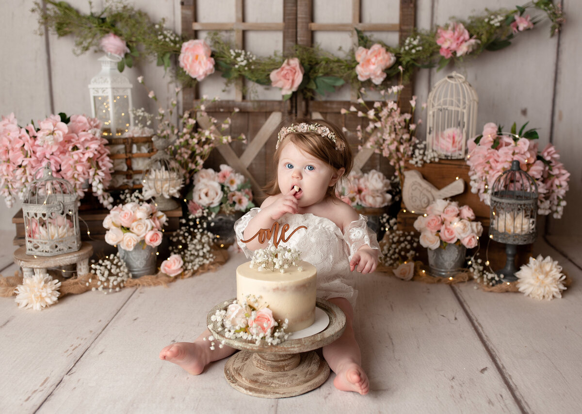 Rose Garden floral themed cake smash at top West Palm Beach, FL photography studio. Baby girl in a white lace romper is licking icing off of her finger. She is sitting behind a cream -colored cake with flowers. In the background, there are rustic bard doors, and several buckets and vases of pink, blush, and cream flowers.