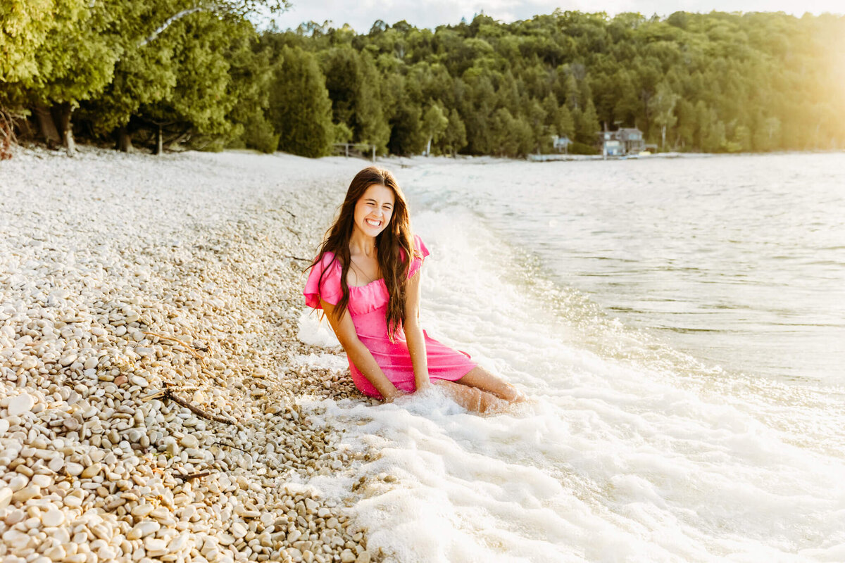 while sitting on the shoreline teen girl in pink dress has wave get her wet during senior photo session