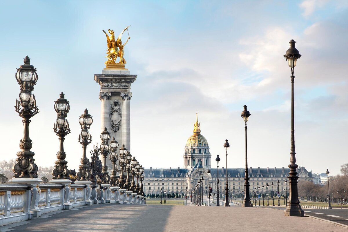 Elegant bridge lined with ornate lampposts leads to gold-topped pillars, featuring a winged statue. In the distance, a grand building with a golden dome is visible under a partly cloudy sky.