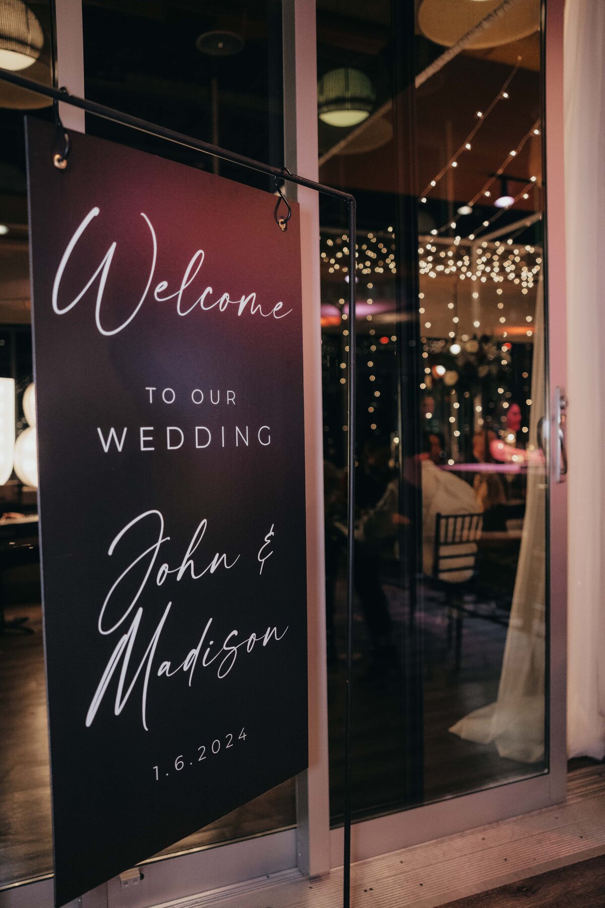 A wedding welcome sign featuring the names "John &amp; Madison" and the date "1.6.2024" at the entrance of a venue with interior lights visible, meticulously arranged by a wedding coordinator