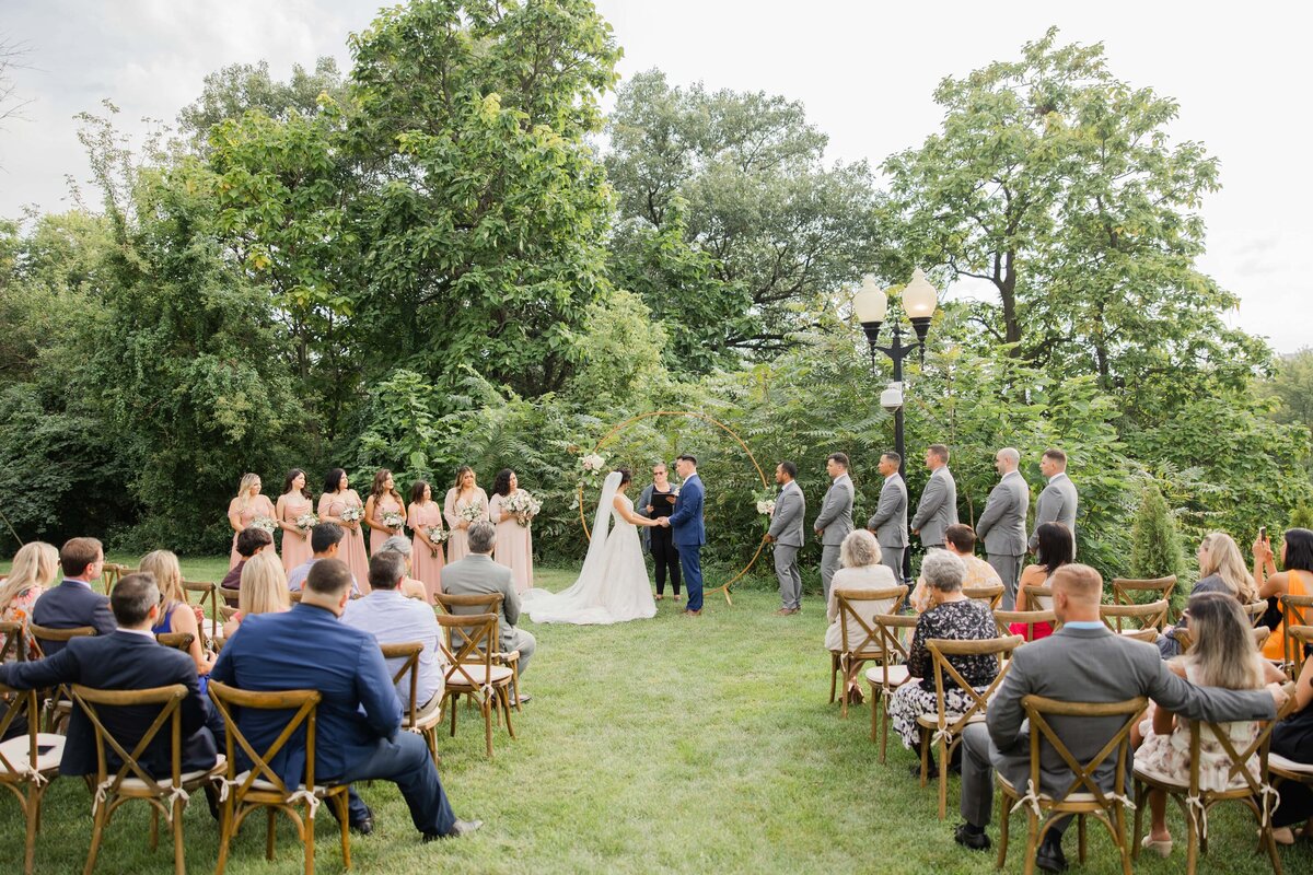 A wedding ceremony in a garden setting at a park farm winery, with a couple exchanging vows under an archway, surrounded by guests and lush greenery.