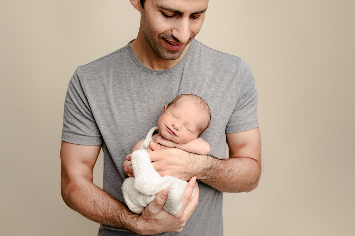 Father looks down towards his newborn son and his hands where you can see how small the baby is compared to Daddy's Hands