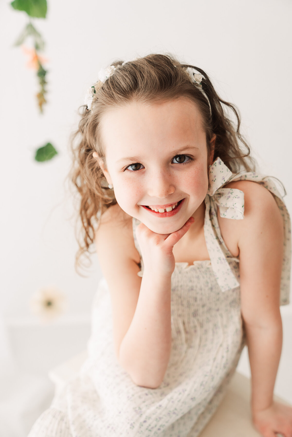 A cheerful young girl with curly hair and a floral hairband smiles brightly while posing in a light, airy room.