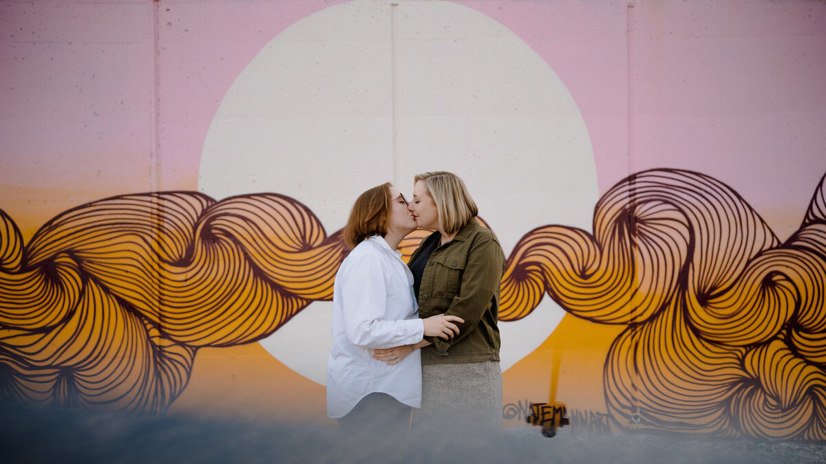 A couple kissing in front of a wall with a mural painted on it.
