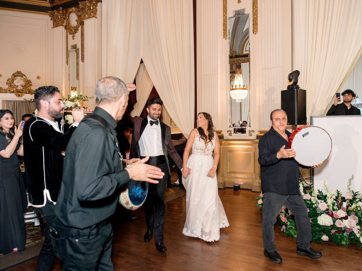 A bride and groom dance joyfully in an elegant ballroom. Musicians play handheld drums, and a man with a microphone entertains the guests. Ornate decorations and flowers adorn the space, creating a festive atmosphere.