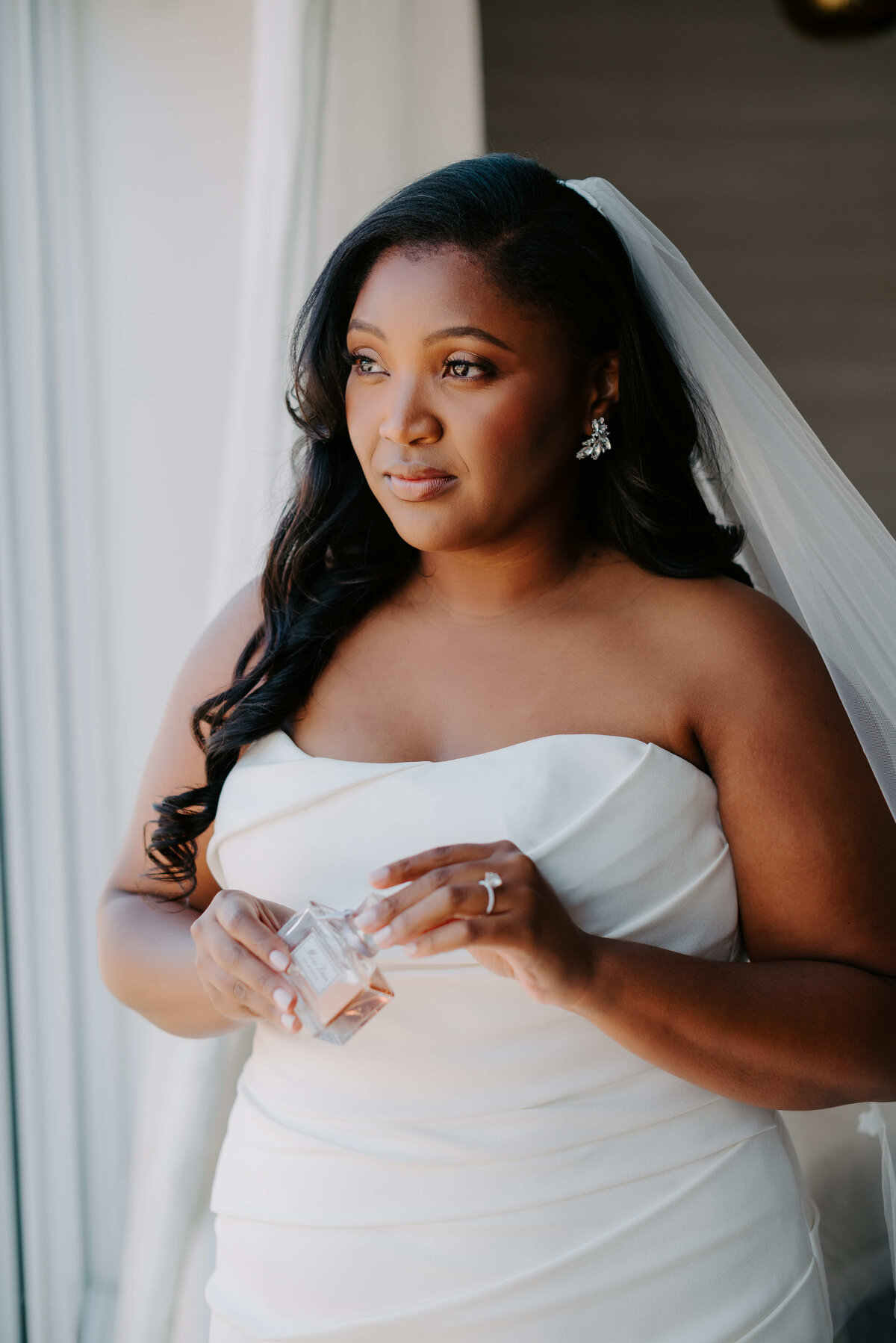A woman in a strapless white wedding dress and veil stands by a window, holding a small bottle and gazing into the distance, beautifully captured by a Dallas wedding photographer.